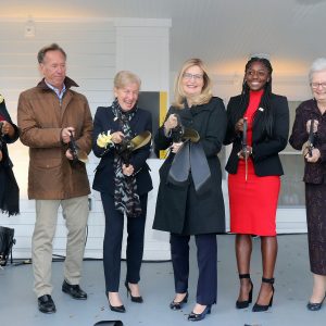 six people cutting a large ribbon with large scissors on the steps of the Michelle Cuozzo Borisenok '80 House
